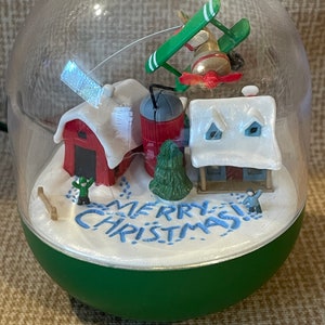 May include: A glass snow globe with a green and red airplane, a red barn, a white house, a green tree, and two figures in the snow. The snow globe has a green base and the words "Merry Christmas!" written in blue on the snow.