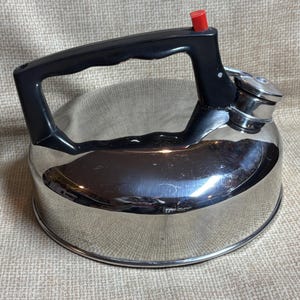 May include: A shiny, stainless steel stovetop kettle with a black handle and a red button on the spout. The kettle has a rounded body and a polished finish, reflecting the surrounding environment.