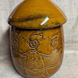 May include: A brown ceramic jar with a lid. The jar features embossed figures of people in festive attire, with snowflake accents. The jar has a glossy finish and a rounded shape, suitable for kitchen storage.