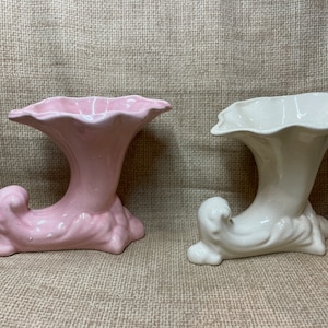 Può includere: Due vasi in ceramica a forma di cornucopia, uno rosa e uno bianco. Entrambi i vasi hanno un'apertura a forma di tromba e una base ricurva.