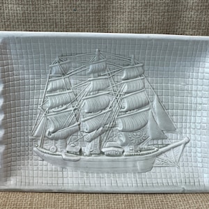 Op de afbeelding: Witte keramische asbak met een reliëfdesign van een zeilschip met meerdere zeilen. De asbak heeft een rasterpatroon en een verhoogde rand.