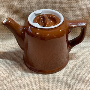 Może przedstawiać: Brązowy ceramiczny czajnik z białą pokrywką i uchwytem. Czajnik ma zaokrąglony korpus i dzióbek.