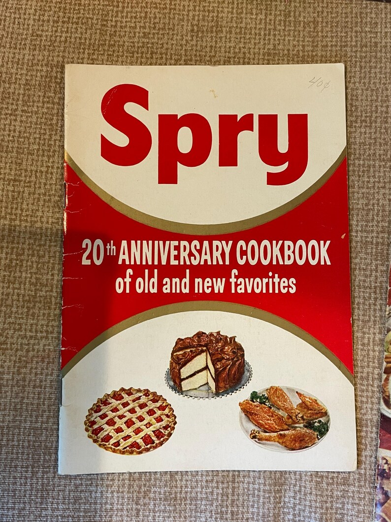 K&ouml;nnte beinhalten: Ein Vintage-Kochbuch mit dem Titel "Spry 20th Anniversary Cookbook of old and new favorites". Der Umschlag zeigt ein rotes und goldenes Design mit Illustrationen von einem Kuchen, einer Torte und Brath&auml;hnchen.