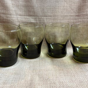 May include: Four clear drinking glasses with a dark green base. The glasses have a rounded shape and are empty. The background is a light brown fabric.