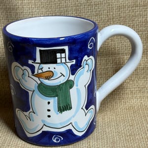 May include: A ceramic mug with a whimsical snowman design. The mug features a white snowman with a black top hat, orange nose, and green scarf against a dark blue background. The handle is white, and the interior is also white.