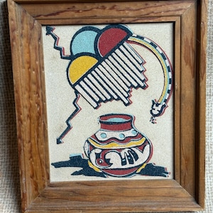 May include: A framed sand art piece with a Native American design. The artwork features a colourful pot, geometric shapes, and a lightning bolt against a beige background. The wooden frame adds a rustic touch.