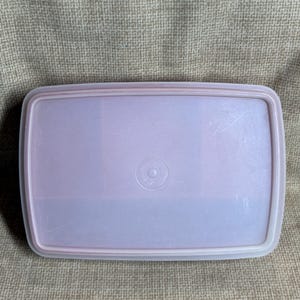 May include: A rectangular, light purple Tupperware lid with a white rim. The lid has a slightly textured surface and a circular emblem in the center. The lid is designed to fit a rectangular container.