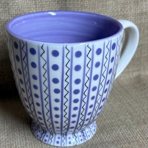May include: A white ceramic mug with a lavender interior and handle. The exterior features vertical stripes of lavender dots and black zigzags. The mug sits on a small, decorative foot.