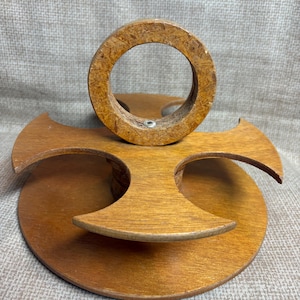 May include: A wooden, multi-tiered display stand with a circular top and a cross-shaped base. The stand is made of light brown wood and has a natural wood grain finish. The circular top is approximately 3 inches in diameter.