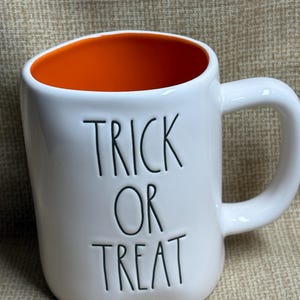 May include: White ceramic mug with an orange interior and a curved handle. The words "TRICK OR TREAT" are printed in black, block letters on the front of the mug. A Halloween-themed mug.