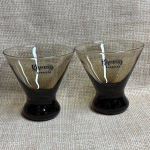 May include: Two Kahlua Special cocktail glasses. The glasses have a dark brown base and a lighter brown bowl. The Kahlua logo is printed on the front of each glass. The glasses are on a neutral-coloured surface.