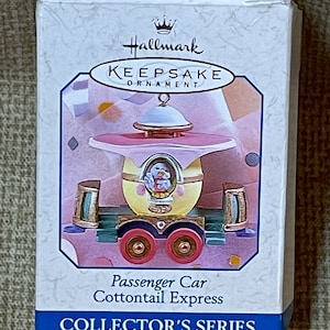 May include: Hallmark Keepsake Ornament, Passenger Car Cottontail Express, Collector's Series. A white box with a gold crown and the words Hallmark Keepsake Ornament. The ornament is a pastel pink, yellow, and blue passenger car with a bunny inside. The car has gold wheels and a gold trim.