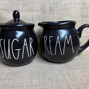May include: Black ceramic sugar bowl with lid and a cream jug. The sugar bowl has the word "SUGAR" in white lettering. The cream jug has the word "CREAM" in white lettering.
