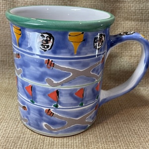 May include: A blue ceramic mug with a green rim and a colourful design of flags, abstract shapes, and lines. The mug has a handle.