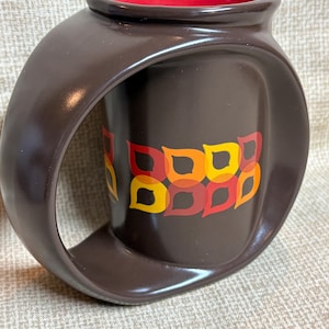 Pode incluir: Uma caneca de cerâmica marrom com interior vermelho e um design de alça exclusivo. A caneca apresenta um padrão retrô de formas de folhas laranja, amarelas e vermelhas. A caneca tem um acabamento brilhante e uma alça curva distinta.
