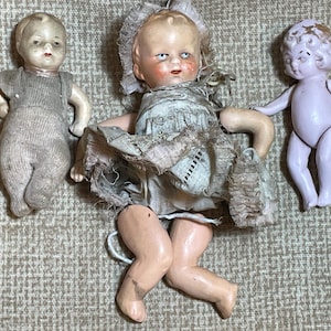May include: Three vintage porcelain dolls. One doll is wearing a white dress with lace trim and has a pink complexion. Another doll is wearing a grey knitted outfit and has a pale complexion. The third doll is wearing a purple outfit and has a pale complexion.