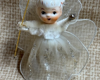 May include: A vintage-style angel figurine with a painted face, white fuzzy bodice, and a sheer, pleated skirt adorned with silver glitter accents. The figure features large, translucent wings, a gold-colored wand, and a small metallic sphere.