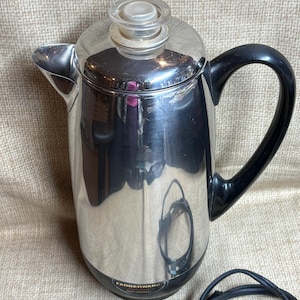 Könnte beinhalten: Ein Edelstahl-Kaffeeperkolator von Farberware mit schwarzem Griff und Knauf. Der Perkolator hat einen Deckel aus klarem Glas und einen Ausguss zum Ausgießen. Auf der Basis befindet sich das Farberware-Logo.