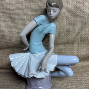 May include: A porcelain figurine of a seated figure with brown hair, wearing a light blue short-sleeved top and a white pleated skirt. The figure is positioned on a light purple tufted base, with light blue leggings visible on the legs.