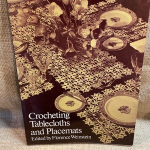 Puede incluir: Un libro titulado "Crocheting Tablecloths and Placemats" editado por Florence Weinstein. La portada presenta una fotografía vintage de una mesa puesta con manteles individuales de ganchillo y un ramo de flores.