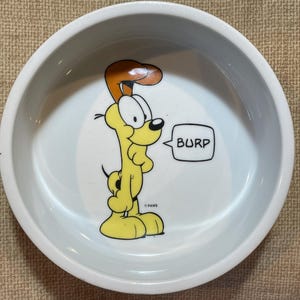 May include: White ceramic pet bowl featuring a cartoon dog character in yellow and orange. The dog is burping, with the word "BURP" in a speech bubble. The bowl has a rounded shape and a wide rim.