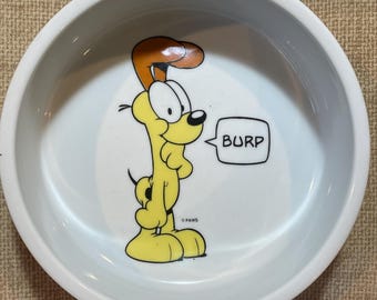 Puede incluir: Cuenco blanco de cerámica para mascotas con un personaje de perro de dibujos animados en amarillo y naranja. El perro eructa, con la palabra "BURP" en un bocadillo. El cuenco tiene forma redondeada y un borde ancho.