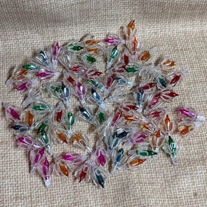 May include: A collection of small, clear Christmas light bulbs with colorful tips. The bulbs have tips in various colors, including red, green, orange, pink, and blue. The bulbs are scattered on a tan fabric surface.