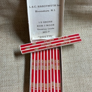 Pode incluir: Uma caixa aberta de grafites de desenho KOH-I-NOOR, com o texto "L. & C. HARDTMUTH Inc." e "Bloomsbury, N. J." A caixa contém lápis vermelhos e brancos marcados "2200 D - 5H." Os lápis estão dispostos em filas.