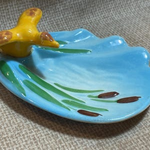 Puede incluir: Un plato de cerámica con forma de concha con un pájaro amarillo posado en el borde. El plato está pintado con un fondo azul, juncos verdes y espadañas marrones.