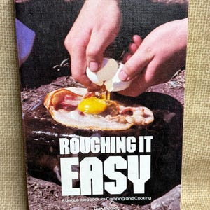 May include: A vintage cookbook titled "Roughing It Easy" with a cover image of hands cracking an egg onto bacon and a fried egg. The book is a unique ideabook for camping and cooking.