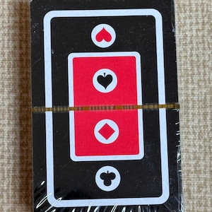 May include: A sealed deck of playing cards with a black background and white borders. The center of the deck shows the suits of hearts, spades, diamonds, and clubs in red and white.