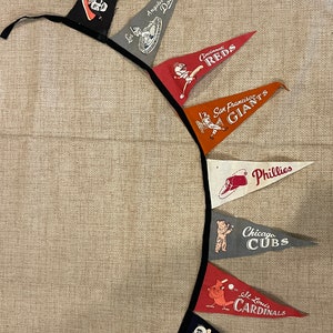 May include: A pennant garland featuring various Major League Baseball team logos. The pennants are in team colors, including black, gray, red, orange, white, and navy. Teams include the Pirates, Dodgers, Reds, Giants, Phillies, Cubs, Cardinals, and Braves.
