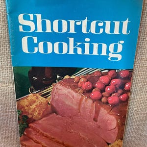 K&ouml;nnte beinhalten: Ein blaues Kochbuch mit dem Titel "Shortcut Cooking" auf dem Cover. Das Cover zeigt ein Foto von geschnittenem Schinken mit einer Kirschso&szlig;e.