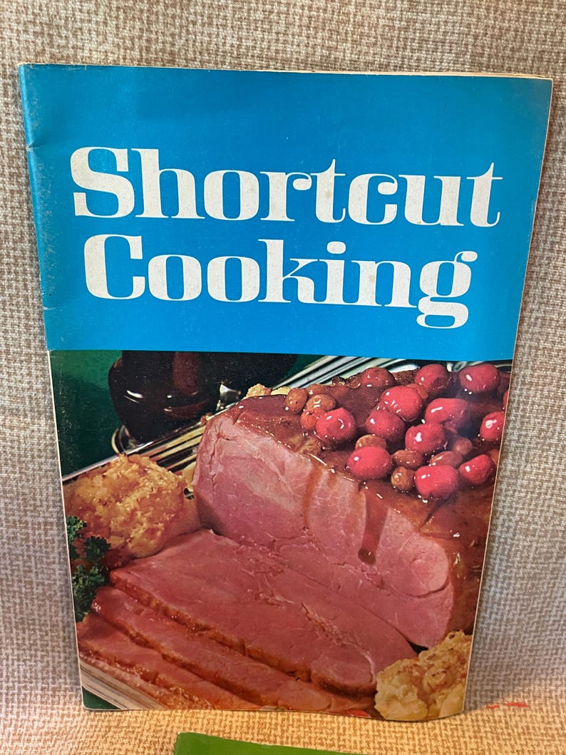 K&ouml;nnte beinhalten: Ein blaues Kochbuch mit dem Titel "Shortcut Cooking" auf dem Cover. Das Cover zeigt ein Foto von geschnittenem Schinken mit einer Kirschso&szlig;e.