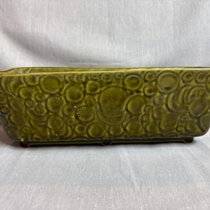 May include: An olive green ceramic planter, rectangular in shape, featuring a textured pattern of circles. The planter has a gently flared form and rests on small feet. This classic design is suitable for a variety of plants and decorative settings.