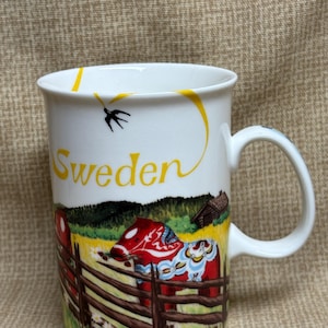 May include: White ceramic mug featuring a colorful illustration of a red Dala horse behind a wooden fence, with the word "Sweden" in yellow. A black bird flies near the rim. The mug has a curved handle.