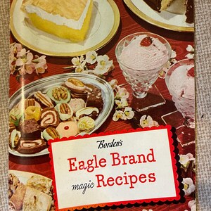 K&ouml;nnte beinhalten: Ein Vintage-Kochbuchcover mit rotem Hintergrund und wei&szlig;en Blumen. Der Titel lautet "Borden's Eagle Brand magic Recipes". Das Cover zeigt auch Bilder von Desserts, darunter einen Zitronen-Baiser-Kuchen, einen Schokoladenkuchen und Eiscreme.