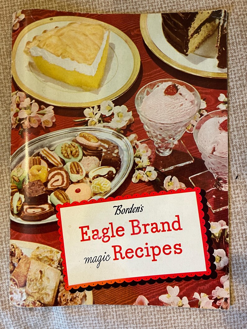 K&ouml;nnte beinhalten: Ein Vintage-Kochbuchcover mit rotem Hintergrund und wei&szlig;en Blumen. Der Titel lautet "Borden's Eagle Brand magic Recipes". Das Cover zeigt auch Bilder von Desserts, darunter einen Zitronen-Baiser-Kuchen, einen Schokoladenkuchen und Eiscreme.