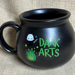 May include: A matte black cauldron-shaped mug featuring a graphic of a small white character holding a wand above green flames. The text "DARK ARTS" is printed in a bright green font to the right of the character, accompanied by small star icons.