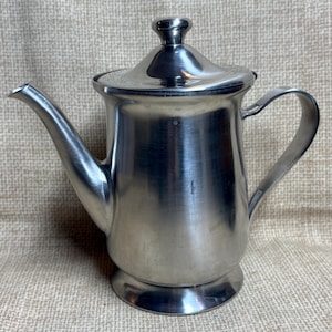 Puede incluir: Una tetera de acero inoxidable con un pico curvo, un asa y una tapa con un pequeño pomo. La tetera tiene una superficie brillante y reflectante y se asienta sobre una base ligeramente elevada. El fondo es una tela texturizada de tonos neutros.