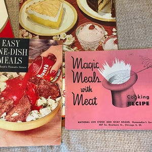 K&ouml;nnte beinhalten: Drei Vintage-Kochb&uuml;cher mit bunten Umschl&auml;gen. Das erste Kochbuch ist rot mit dem Text "Easy One-Dish Meals with Hunt's Tomato Sauce". Das zweite Kochbuch ist rosa mit dem Text "Magic Meals with Meat" und einer Grafik eines Zylinders mit einer wei&szlig;en Wolke, die aus ihm herauskommt. Das dritte Kochbuch ist rosa mit dem Text "Cooking School Recipe Book" und einer Grafik einer Hand, die einen Zauberstab h&auml;lt.