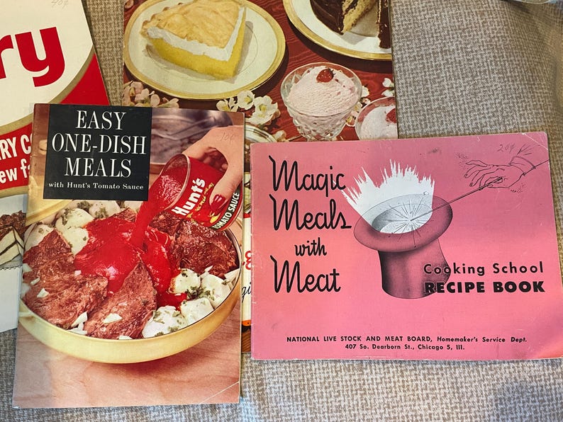 K&ouml;nnte beinhalten: Drei Vintage-Kochb&uuml;cher mit bunten Umschl&auml;gen. Das erste Kochbuch ist rot mit dem Text "Easy One-Dish Meals with Hunt's Tomato Sauce". Das zweite Kochbuch ist rosa mit dem Text "Magic Meals with Meat" und einer Grafik eines Zylinders mit einer wei&szlig;en Wolke, die aus ihm herauskommt. Das dritte Kochbuch ist rosa mit dem Text "Cooking School Recipe Book" und einer Grafik einer Hand, die einen Zauberstab h&auml;lt.