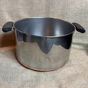 以下が含まれることがあります： 銅色の底と2つの黒いハンドルが付いたステンレス製の調理鍋。鍋は光沢のある反射面を持ち、調理用に設計されています。食品の沸騰や煮込みに使用される可能性があります。