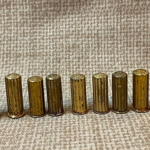 Puede incluir: Nueve casquillos de metal de color dorado, posiblemente para balas o cartuchos, están alineados en una fila sobre una superficie marrón texturizada.