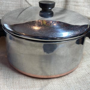 May include: A stainless steel cooking pot with a lid and black handles. The pot has a copper-coloured base and a black knob on the lid. The pot is on a beige surface.