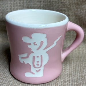 May include: Pink ceramic mug with a white interior and a white cartoon figure design. The mug has a curved handle and a rounded base. The cartoon figure is wearing a hat and appears to be dancing.
