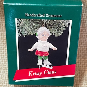 Peut inclure: Une boîte verte et blanche avec le logo Hallmark et les mots "Ornement artisanal" sur le devant. La boîte contient une petite figurine en porcelaine blanche d'une fille portant un chapeau vert, un pull blanc, une jupe rouge et un pantalon vert. La figurine tient une paire de patins à glace argentés et porte les mots "Kristy Claus" sur son pull. La boîte porte également les mots "Kristy Claus" sur le devant.