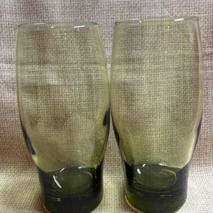 Peut inclure: Deux verres à boire teintés vert olive. Les verres ont une forme cylindrique avec une base vert foncé. L'arrière-plan est un tissu texturé de ton neutre.