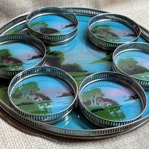 以下が含まれることがあります： 6つのコースターとトレイのヴィンテージセット。トレイとコースターは銀色の金属リム付き。各コースターとトレイの表面には、湖、木、家のある風景デザインが施されています。トレイの直径は約30cmです。