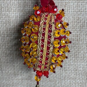 May include: A hanging ornament featuring a deep red fabric base decorated with gold metallic trim, yellow faceted beads, and small red beads. The ornament is suspended by a red velvet ribbon and includes a small teardrop-shaped charm at the bottom.